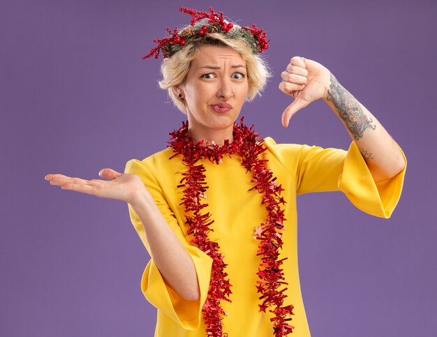 Unpleased young blonde woman wearing christmas head wreath and tinsel garland around neck looking at camera showing empty hand and thumb down isolated on purple background