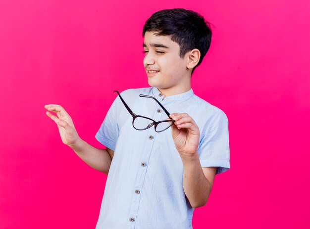 Unpleased young boy holding glasses keeping hand in air with closed eyes gesturing no isolated on pink wall with copy space