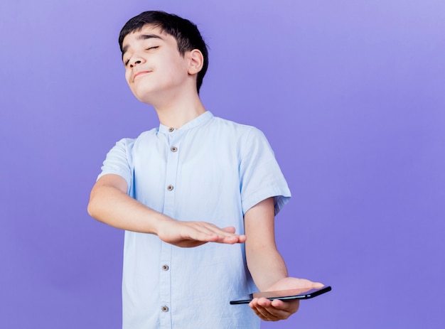 Unpleased young boy holding mobile phone doing no gesture with hand with closed eyes isolated on purple wall