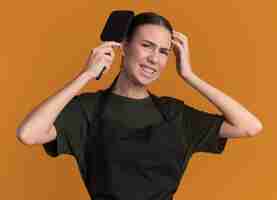 Free photo unpleased young brunette barber girl in uniform combing hair  isolated on orange wall with copy space