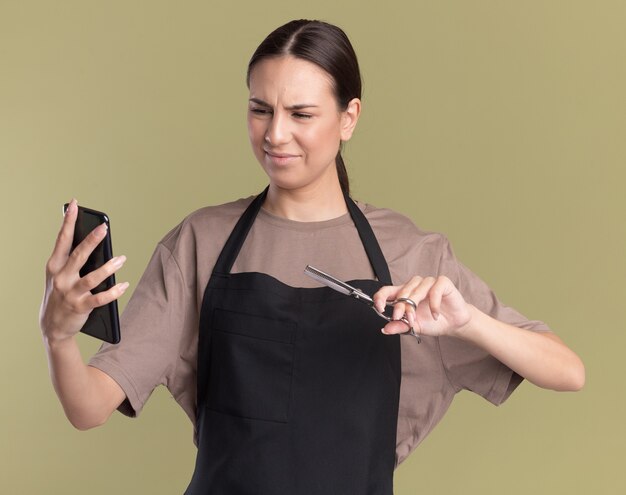 Unpleased young brunette barber girl in uniform holds hair thinning scissors and looks at phone isolated on olive green wall with copy space