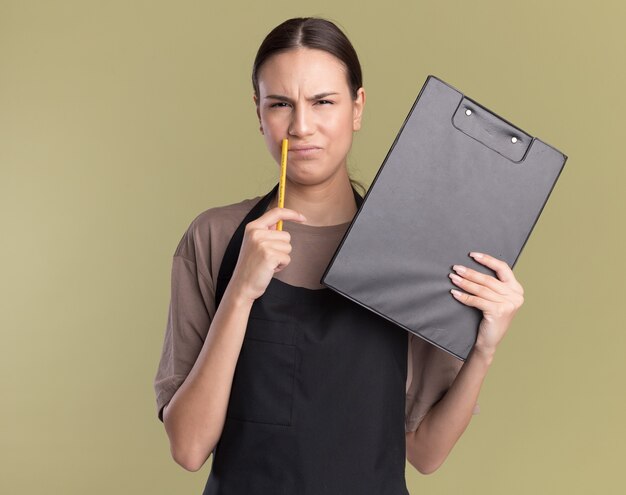 Unpleased young brunette barber girl in uniform holds pencil and clipboard on olive green