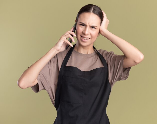 Unpleased young brunette barber girl in uniform puts hand on head and talks on phone on olive green