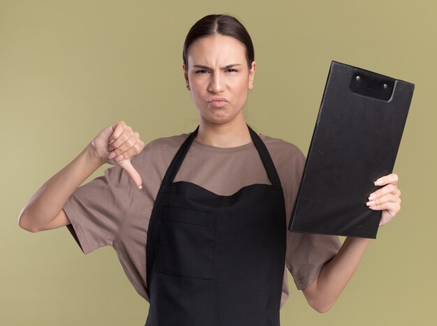 Unpleased young brunette barber girl in uniform thumbs down and holds clipboard isolated on olive green wall with copy space