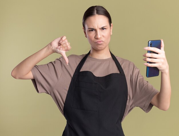 Unpleased young brunette barber girl in uniform thumbs down and holds phone on olive green