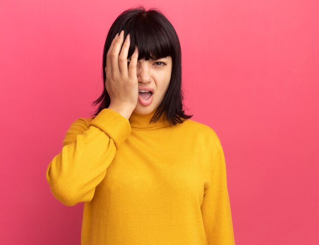 Unpleased young brunette caucasian girl covers eye with hand on pink