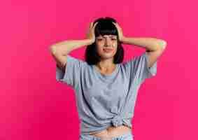 Free photo unpleased young brunette caucasian girl holds head looking at camera isolated on pink background with copy space