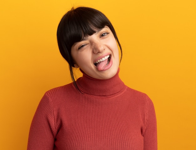 Unpleased young brunette caucasian girl stucks out tongue isolated on orange wall with copy space