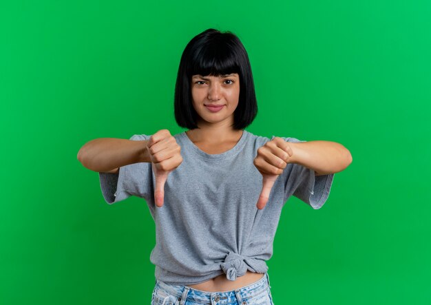 Unpleased young brunette caucasian girl thumbs down with two hands isolated on green background with copy space