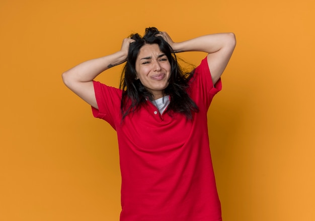 Unpleased young brunette caucasian girl wearing red shirt holds head isolated on orange wall