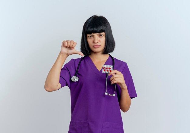 Unpleased young brunette female doctor in uniform with stethoscope holds and points at medicine 