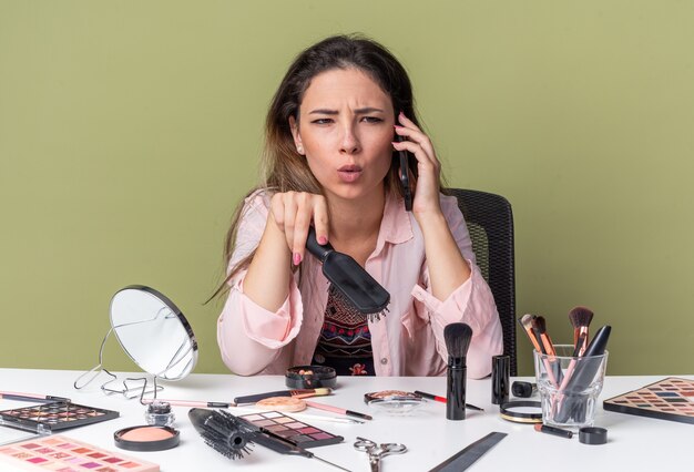 Unpleased young brunette girl sitting at table with makeup tools talking on phone and holding comb 