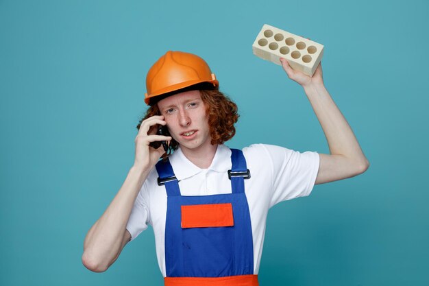 Unpleased young builder man in uniform holding brick and speak on the phone isolated on blue background
