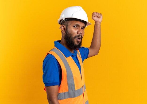 안전 헬멧을 쓴 제복을 입은 불쾌한 젊은 건축업자가 복사 공간이 있는 주황색 벽에 고립되어 주먹을 들고 있다