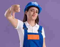 Бесплатное фото Недовольная молодая женщина-строитель в униформе показывает большой палец вниз изолированной на фиолетовой стене