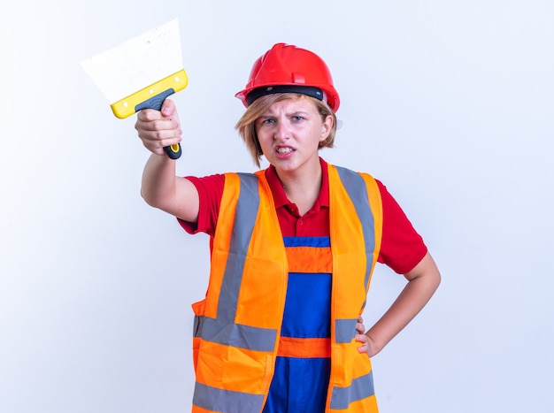 unpleased young builder woman in uniform holding out putty knife at front isolated on white wall