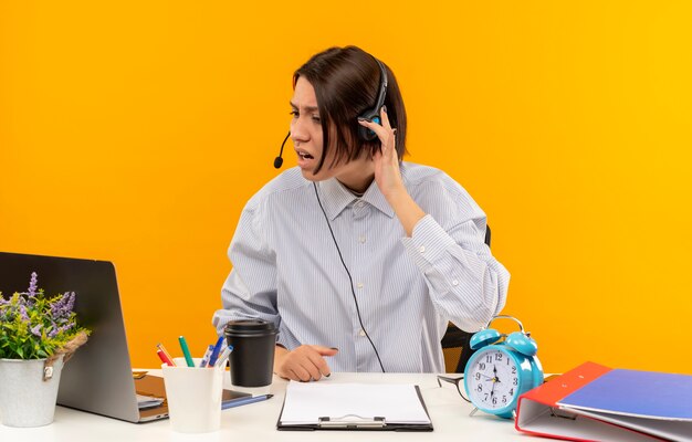 Unpleased young call center girl wearing headset sitting at desk with work tools looking at laptop with hand on headset isolated on orange background