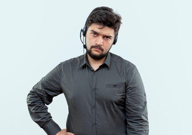 Unpleased young call center man wearing headset putting hand on hip isolated on white