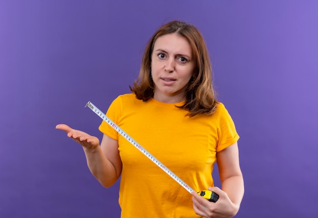 Unpleased young casual woman holding tape meter and showing empty hand on isolated purple wall
