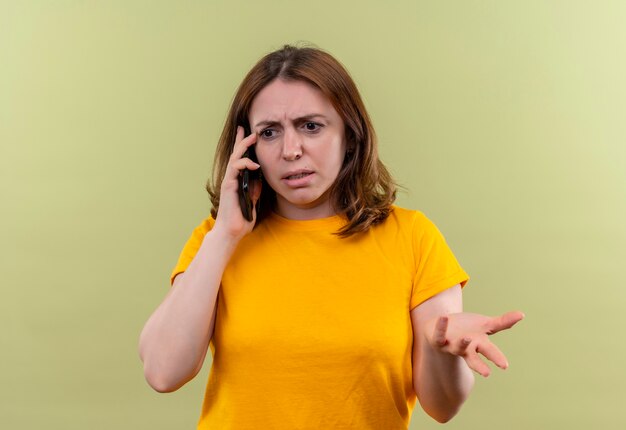 Unpleased young casual woman talking on phone and showing empty hand on isolated green wall
