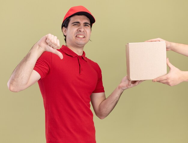 Unpleased young caucasian delivery man in red uniform and cap giving cardbox to client showing thumb down 