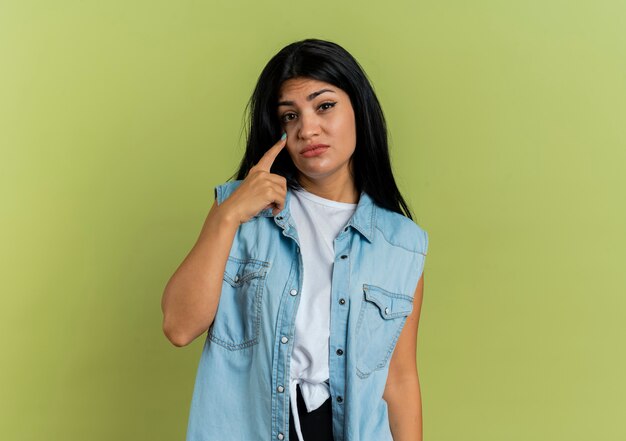 Unpleased young caucasian girl pulling down eyelid isolated on olive green background with copy space