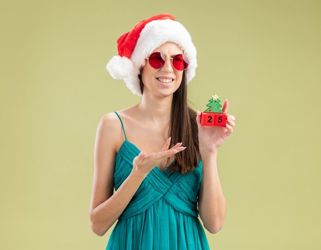 Unpleased young caucasian girl in sun glasses with santa hat holding and pointing at christmas tree ornament with hand 