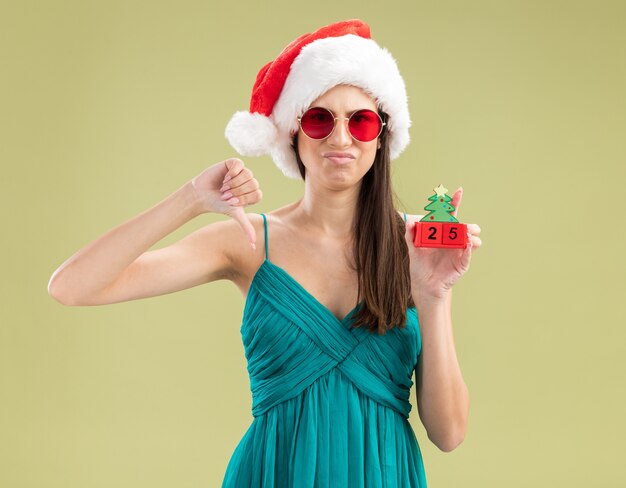 산타 모자와 태양 안경에 불쾌한 젊은 백인 여자는 크리스마스 트리 장식을 보유하고 아래로 엄지 손가락