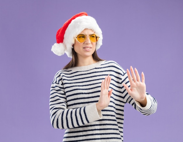 Free photo unpleased young caucasian girl in sun glasses with santa hat holds hands open gesturing no sign looking at side