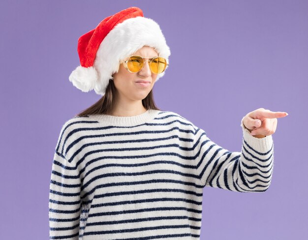 Unpleased young caucasian girl in sun glasses with santa hat looking and pointing at side 