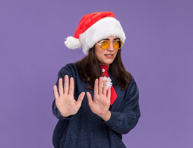 Unpleased young caucasian girl in sun glasses with santa hat and santa tie gesturing stop sign with hands isolated on purple wall with copy space