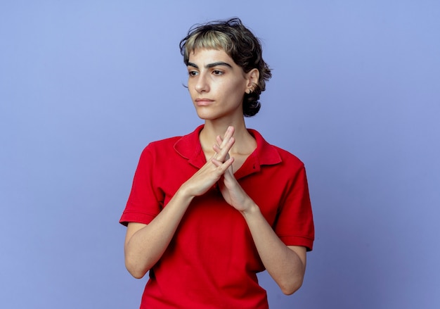 Unpleased young caucasian girl with pixie haircut putting hands together looking at side isolated on purple background with copy space