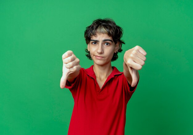 Unpleased young caucasian girl with pixie haircut showing thumbs down isolated on green background with copy space