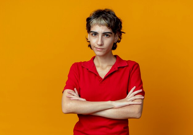 Unpleased young caucasian girl with pixie haircut standing with closed posture isolated on orange background with copy space