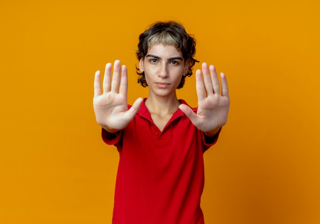 Unpleased young caucasian girl with pixie haircut stretching out hands gesturing stop isolated on orange background with copy space