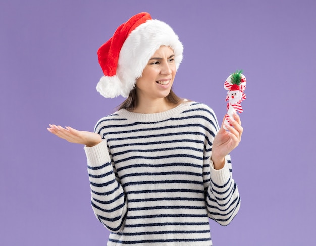 Unpleased young caucasian girl with santa hat holding and looking at candy cane 