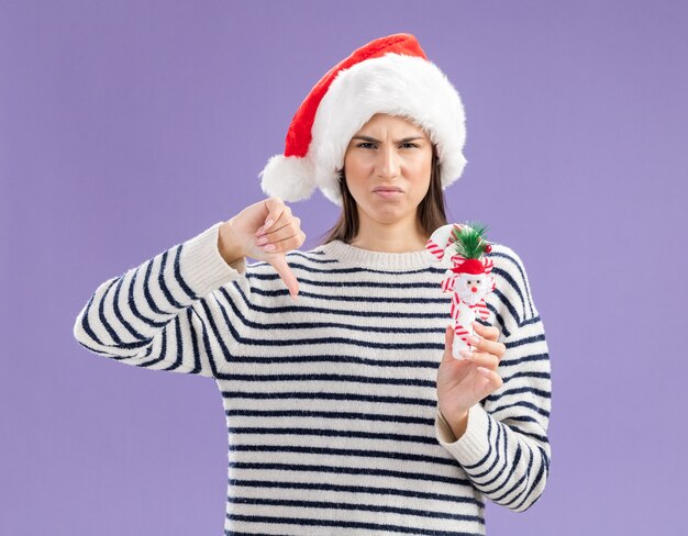 サンタの帽子をかぶった不機嫌な若い白人の女の子は、キャンディケインと親指を押し下げます