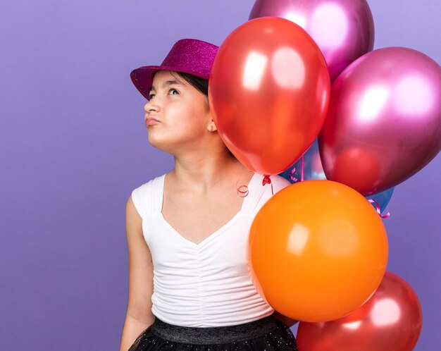 unpleased young caucasian girl with violet party hat holding helium balloons and looking up isolated on purple wall with copy space