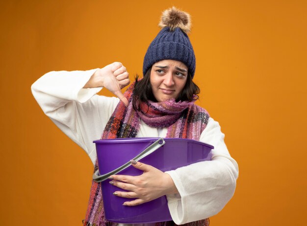 ローブの冬の帽子とスカーフを身に着けている不機嫌な若い白人の病気の女の子は、コピースペースのあるオレンジ色の壁に隔離された親指を下に向けてプラスチック製のバケツを見て吐き気を持っています