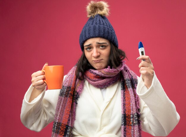 Unpleased young caucasian ill girl wearing robe winter hat and scarf holding cup of tea and thermometer looking at camera isolated on crimson background