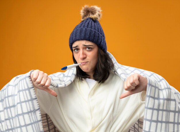 Unpleased young caucasian ill girl wearing robe winter hat wrapped in plaid looking at camera showing thumbs down with thermometer in mouth isolated on orange background