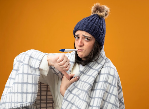 Unpleased young caucasian ill girl wearing robe winter hat wrapped in plaid looking at camera with thermometer in mouth showing thumb down isolated on orange background