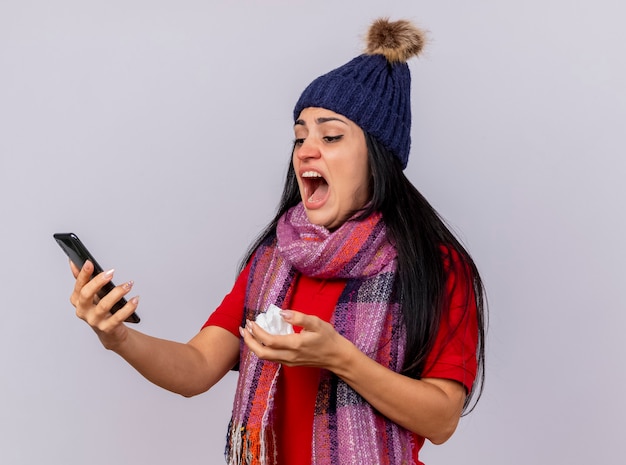 Unpleased young caucasian ill girl wearing winter hat and scarf holding and looking at mobile phone with napkin in hand isolated on white background with copy space