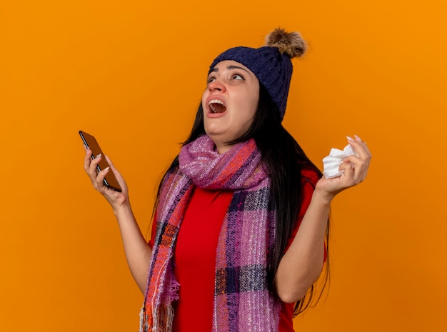 Unpleased young caucasian ill girl wearing winter hat and scarf holding mobile phone and napkin looking up isolated on orange wall