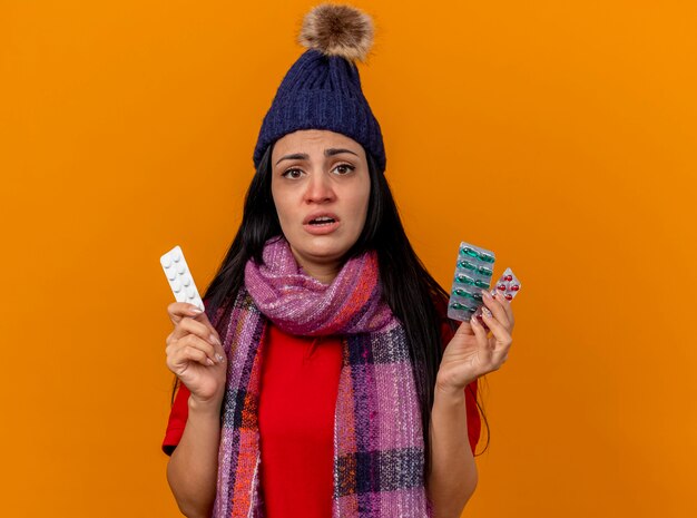 Unpleased young caucasian ill girl wearing winter hat and scarf  holding packs of medical pills isolated on orange wall with copy space