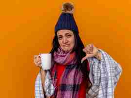 Foto gratuita giovane ragazza malata caucasica dispiaciuta che indossa cappello invernale e sciarpa avvolti in una tazza di tè che tiene plaid che mostra il pollice verso il basso isolato sulla parete arancione con lo spazio della copia