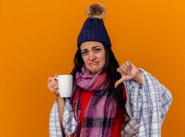 Unpleased young caucasian ill girl wearing winter hat and scarf wrapped in plaid holding cup of tea  showing thumb down isolated on orange wall with copy space