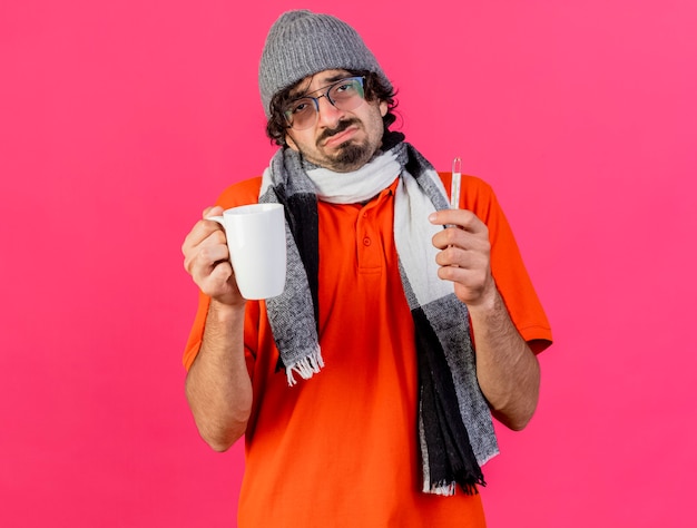 Unpleased young caucasian ill man wearing glasses winter hat and scarf holding cup and thermometer looking at camera isolated on crimson background with copy space