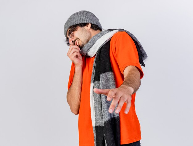 Unpleased young caucasian ill man wearing glasses winter hat and scarf looking at side stretching out hand towards camera keeping another one near mouth isolated on white background with copy space