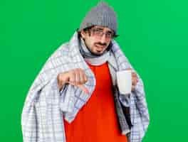 Free photo unpleased young caucasian ill man wearing glasses winter hat and scarf wrapped in plaid holding cup looking at camera showing thumb down isolated on green background with copy space